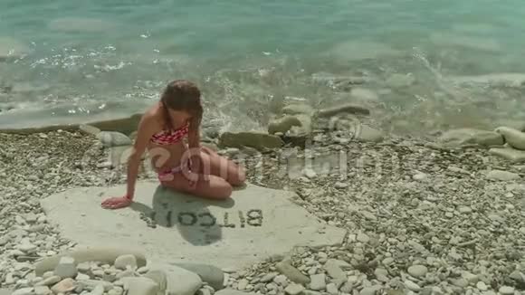 年轻的金发女孩穿着泳衣独自坐在岩石海滩上编造了比特币这个词视频的预览图