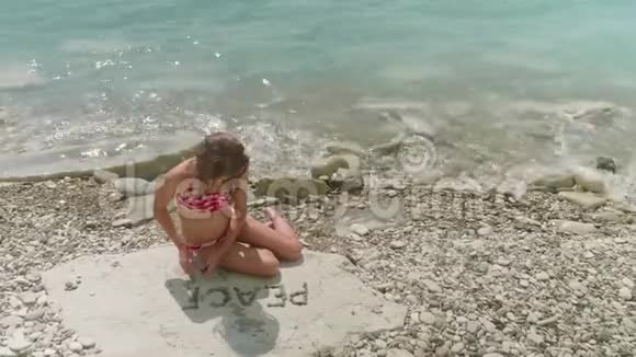年轻的金发女孩穿着泳衣独自坐在岩石海滩上组成了和平这个词视频的预览图