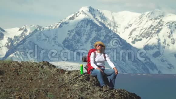 旅行者坐在山顶的女人从热水瓶里喝热茶红色背包橙色夹克草帽筷子视频的预览图