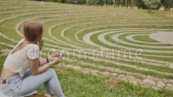 一个年轻的女学生坐在草地上的体育场边缘手里拿着一部手机休息时间视频的预览图
