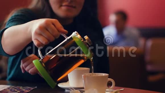 一个年轻的女人慢慢地从玻璃茶壶里把热茶倒进杯子里视频的预览图