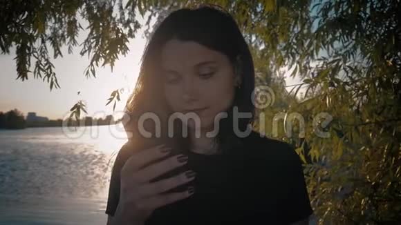 一位年轻漂亮的黑发女人在湖边的夕阳背景下使用智能手机视频的预览图