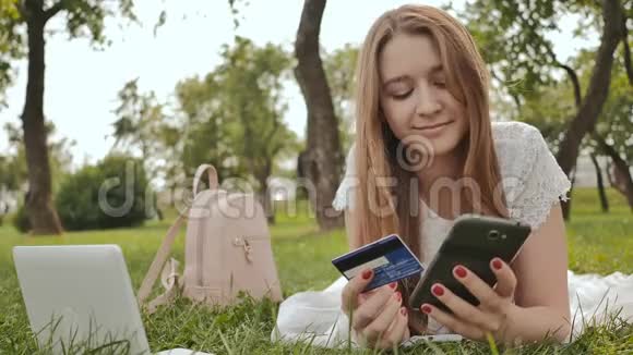 躺在大自然中的一位年轻女孩用笔记本电脑用信用卡通过互联网购物视频的预览图