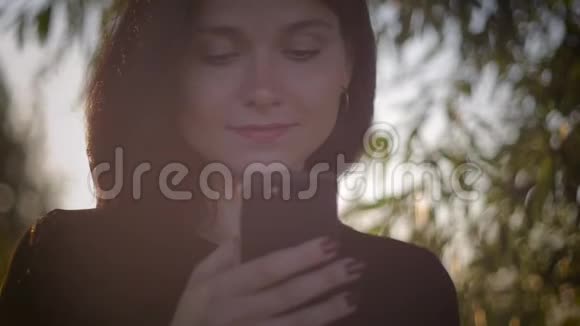 一位年轻漂亮的黑发女人在湖边的夕阳背景下使用智能手机视频的预览图