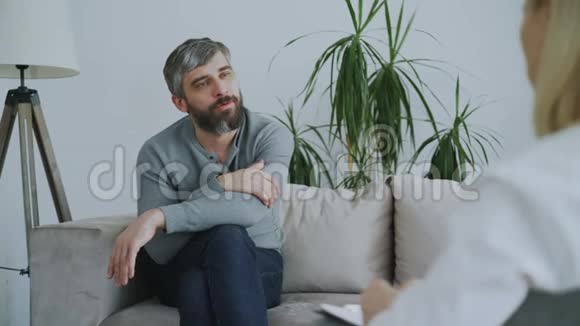 坐在沙发上与办公室内的女心理治疗师交谈的成年长胡子男子视频的预览图