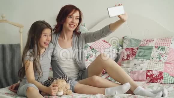 快乐的妈妈和小女孩用智能手机相机拍照坐在舒适的床上做鬼脸视频的预览图