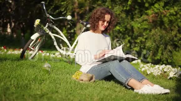 穿着白色衬衫和蓝色牛仔裤的漂亮女人正在阳光明媚的温暖日子里坐在公园的草地上看书她是视频的预览图