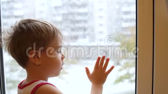 一个孩子站在窗户附近看着雪落在街上玻璃窗上的特写视频的预览图