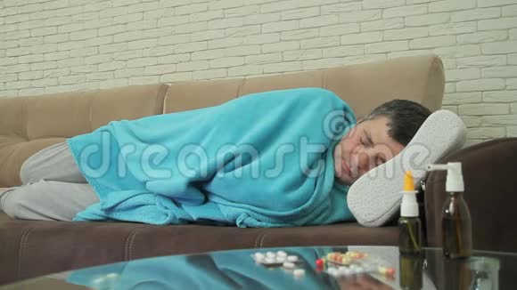 一个有感冒症状的男人躺在沙发上用毯子盖住房间测量房间的温度视频的预览图
