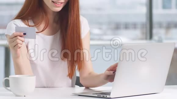 漂亮的红发女孩拿着信用卡在笔记本电脑上进行网上购物视频的预览图