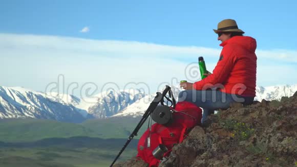 旅行者坐在山顶的女人从热水瓶里喝热茶红色背包橙色夹克草帽筷子视频的预览图