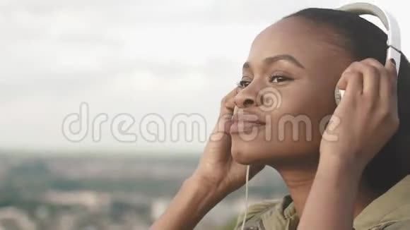 欢快迷人的非洲女人微笑的肖像戴上耳机在模糊的地方欣赏音乐视频的预览图