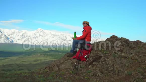 旅行者坐在山顶的女人从热水瓶里喝热茶红色背包橙色夹克草帽筷子视频的预览图
