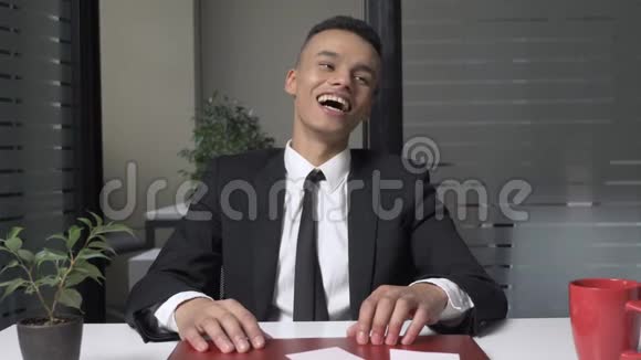 一个穿西装的年轻成功的非洲商人正坐在办公室里一边看着摄像机一边笑视频的预览图