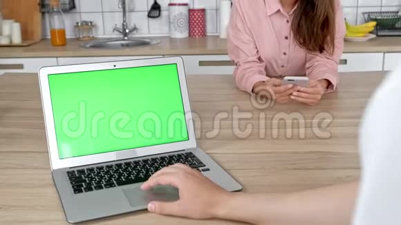 快乐的年轻夫妇在早上一起吃早餐之前在厨房里使用笔记本电脑微笑的男人和女人视频的预览图