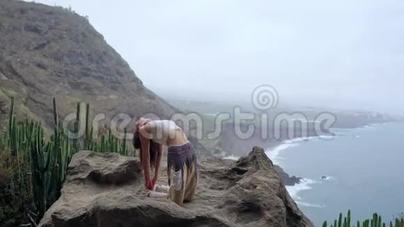 女孩在岩石上练习瑜伽对抗蓝天和蔚蓝的海洋女人站在石头上站在桥上的姿势视频的预览图
