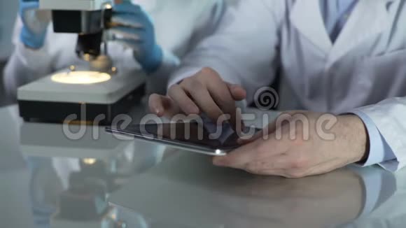 男科学家滚动平板屏幕同事在附近的显微镜工作视频的预览图