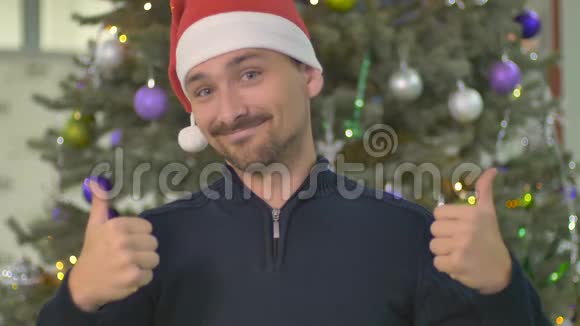圣诞老人帽子里个滑稽男人在背景上用圣诞树竖起大拇指视频的预览图