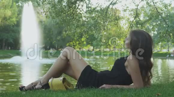 躺在灰色草地上的迷人的黑发女郎在池塘附近放松视频的预览图