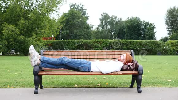 坐在长凳上的公园里穿着红胡子和头发睡累了的人视频的预览图
