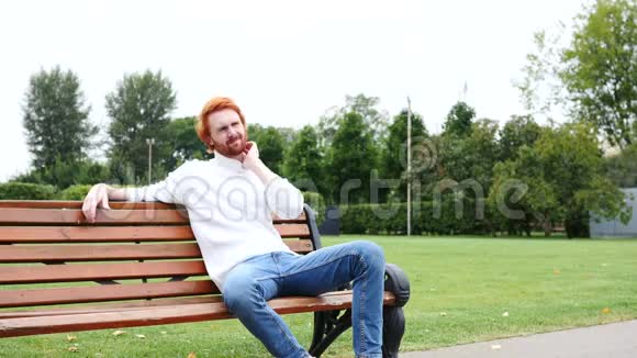 累了脖子痛红胡子和头发坐在公园的长凳上视频的预览图