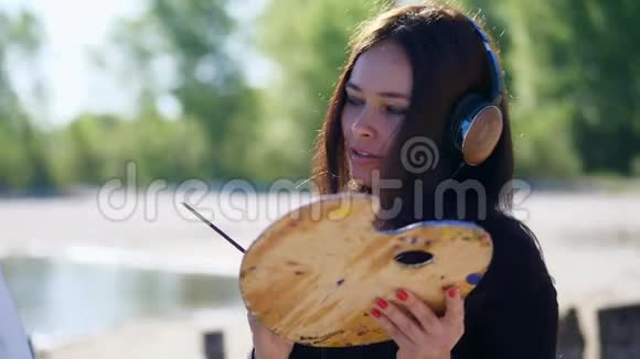 夏天河边海滩上日出时一位戴着棕色大耳机的美女艺术家手里拿着画笔调色板视频的预览图