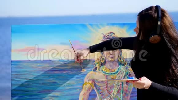 夏天河边海滩上日出时一位戴着棕色大耳机的美女艺术家手里拿着画笔调色板视频的预览图