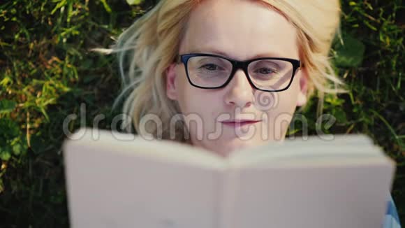 一个戴眼镜的年轻女人的肖像躺在公园的草坪上看书视频的预览图