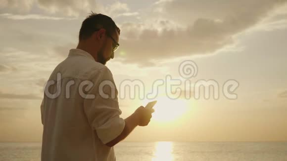 一个在海边度假的年轻人日落时分使用智能手机视频的预览图