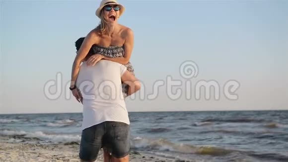 强壮的男人捡起他的女朋友在肩膀上在海上旋转背景穿着长裙的女人笑视频的预览图
