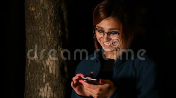 女孩晚上在树附近使用智能手机视频的预览图