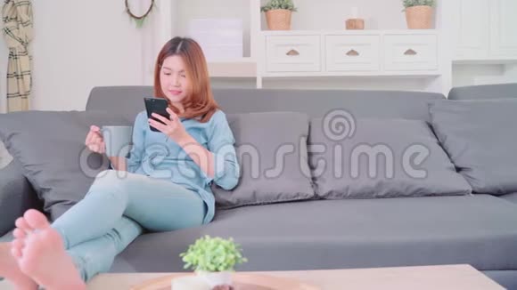 美丽迷人微笑的亚洲女人的肖像她用智能手机拿着一杯温暖的咖啡或茶躺在沙发上视频的预览图