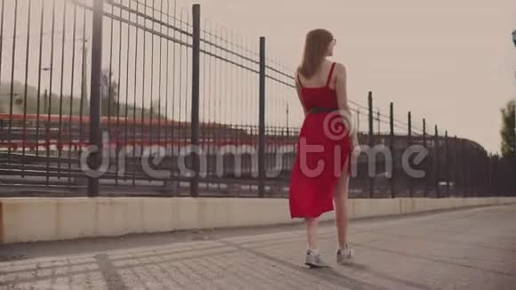 迷人的年轻红发女人穿着红色的裙子走在街上在日落时玩得很开心慢动作视频的预览图