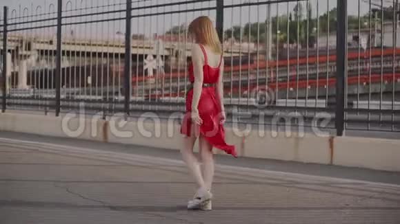迷人的年轻红发女人穿着红色的裙子走在街上在日落时玩得很开心慢动作视频的预览图