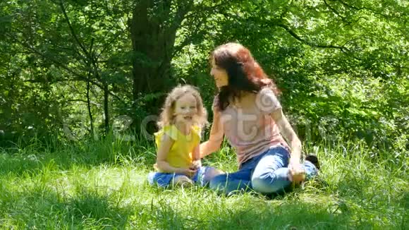有魅力的年轻母亲和小女儿一起坐在城市的绿草上的画像视频的预览图