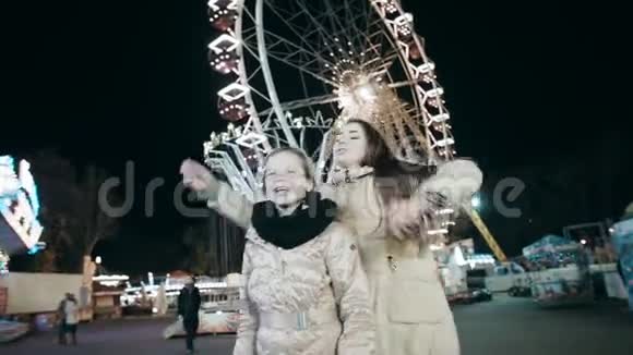两个美丽的年轻女子晚上在一个闪闪发光的摩天轮的背景下的装饰公园跳舞视频的预览图