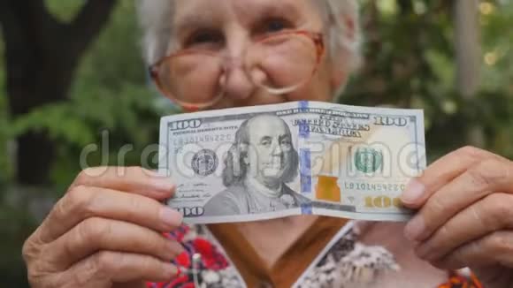 戴眼镜的老太太把一百美元的钞票照进镜头在户外微笑奶奶在外面拿现金视频的预览图