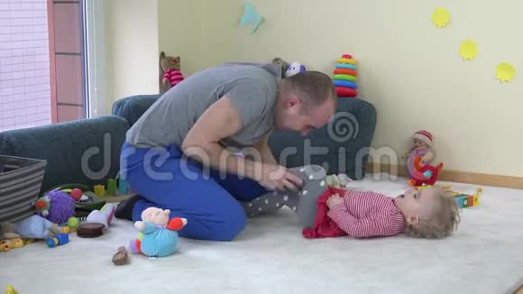快乐的年轻父亲在家玩具之间的地毯上挠着蹒跚学步的女儿视频的预览图