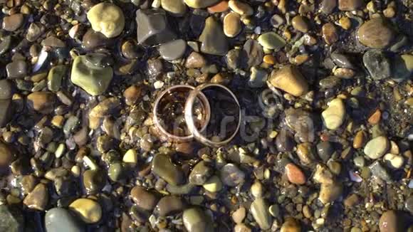 两个金色的结婚戒指躺在沙滩上的石头上阳光照耀着接近宏观环上的光的灌注海洋视频的预览图
