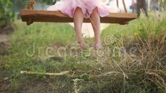 一个穿着粉红色裙子的女孩在农村自制的秋千上溜冰光脚草和土的特写节日快乐视频的预览图
