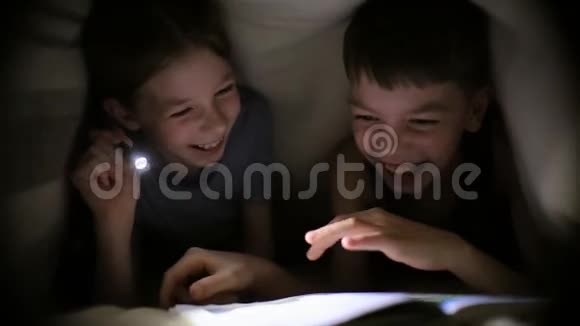 哥哥姐姐晚上在黑暗的房间里用手电筒在毯子下读一本书孩子们在玩视频的预览图