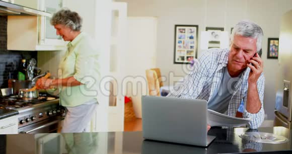 年长男子使用笔记本电脑而妇女则在4k背景下做饭视频的预览图