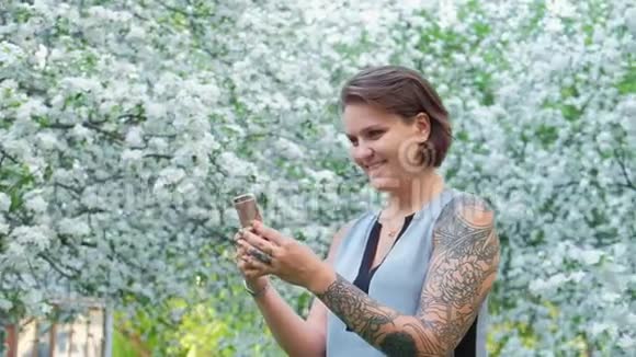 可爱的年轻棕色头发的女人正在阳光明媚的日子里在盛开的苹果树花园里和智能手机聊天生活方式视频的预览图
