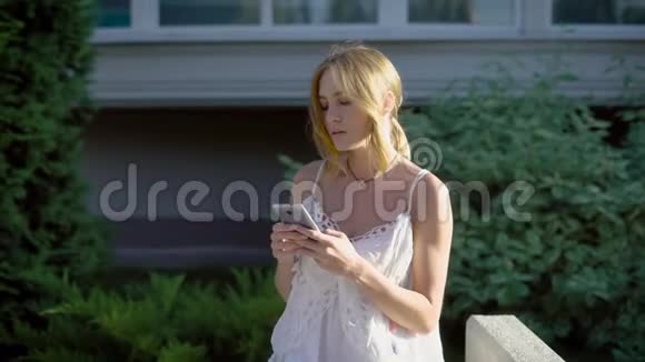 美丽美丽的金发年轻女子正静静地在城市街道上的智能手机上聊天视频的预览图