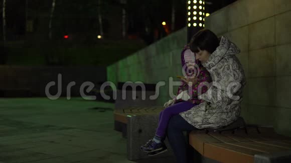 妈妈和女儿坐在长凳上晚上在公园里用电话视频的预览图