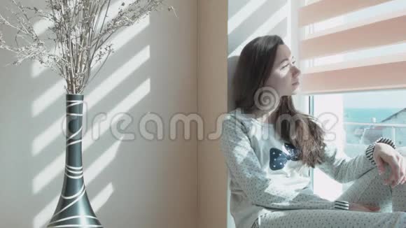 一个年轻漂亮的女人坐在俯瞰大海的窗户上桌子上有一个花瓶上面有装饰花光线视频的预览图