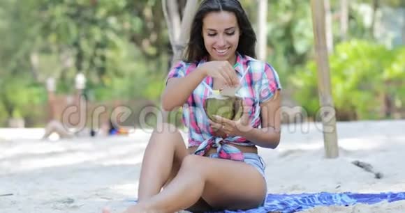 美丽的女孩喝椰子鸡尾酒坐在棕榈树上的海滩上快乐的微笑年轻女性游客视频的预览图