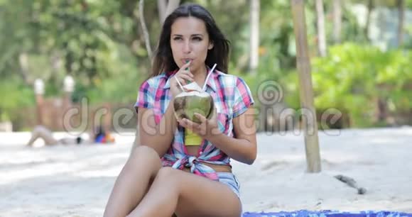 美丽的女孩喝椰子鸡尾酒坐在棕榈树上的海滩上快乐的微笑年轻女性游客视频的预览图