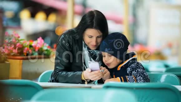一位年轻的母亲带着智能手机在咖啡馆的一张桌子上展示了她可爱的小儿子的一些东西视频的预览图