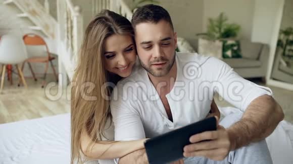 年轻可爱的一对情侣早上坐在卧室的床上使用平板电脑和亲吻视频的预览图